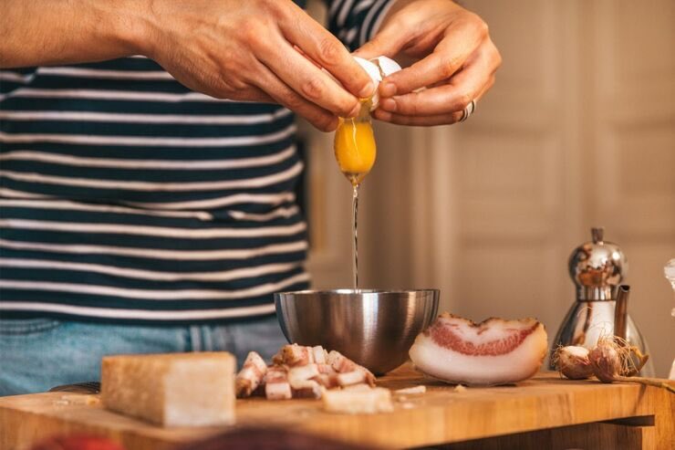 Mani che aprono uovo in una ciotola, guanciale sul tagliere